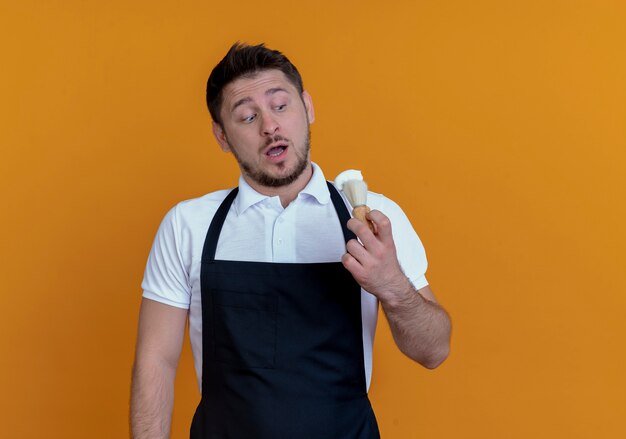 Barber man in apron holding shaving brush looking at it surprised standing over orange background