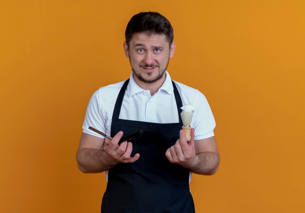 barber man in apron holding shaving brush and razor  with smile on face standing over orange wall