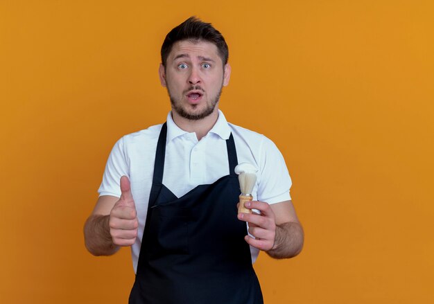 Barber man in apron holding shaving brush showing thumbs up looking at camera confused and surprised standing over orange background