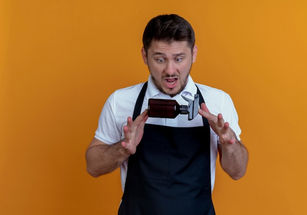 barber man in apron holding spray looking at it surprised and amazed standing over orange wall