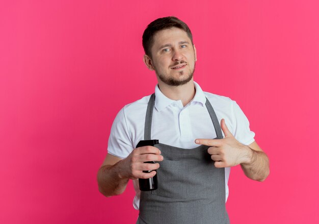 Barber man in apron holding spray pointing with finger to it smiling standing over pink background