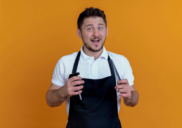 Barber man in apron holding spray and scissors looking at camera smiling with happy face standing over orange background