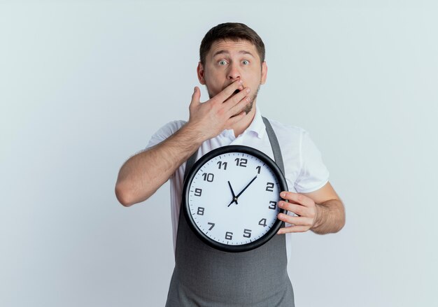 barber man in apron holding wall clock  covering mouth with hand being shocked standing over white wall