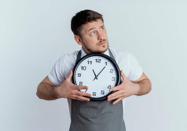 Barber man in apron holding wall clock looking aside puzzled standing over white wall