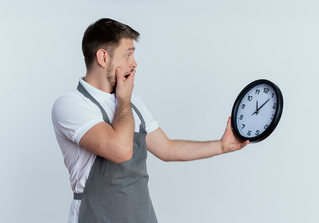 barber man in apron holding wall clock looking at it confused and surprised standing over white wall