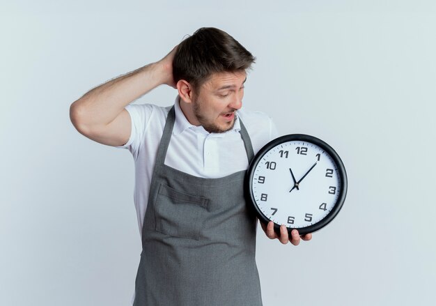 barber man in apron holding wall clock looking at it confused with hand on his head standing over white wall