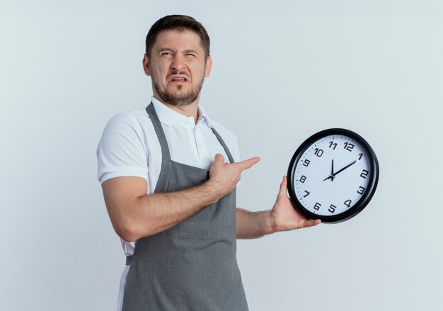 Barber man in apron holding wall clock pointing with finger to it looking confused and very anxious standing over white background