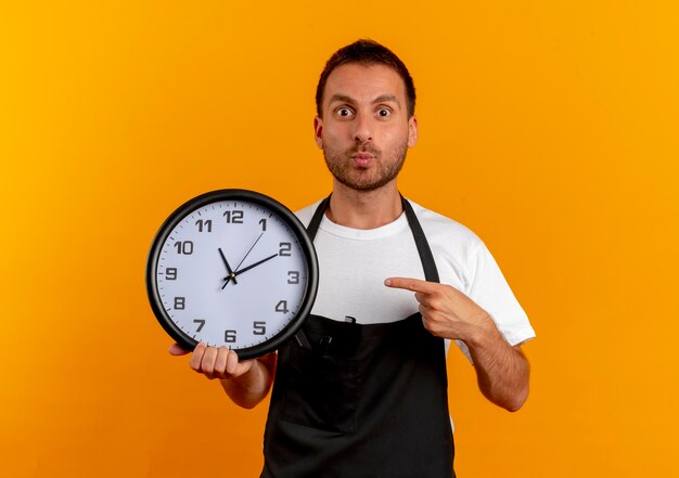 Мужчина-парикмахер в фартуке держит настенные часы, указывая пальцем на него, выглядит удивленным, стоя над оранжевой стеной