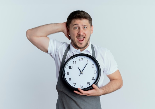 Free photo barber man in apron holding wall clock  puzzled and confused standing over white wall