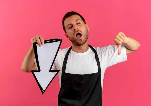 Free photo barber man in apron holding white arrow looking to the front displeased showing thumbs down standing over pink wall