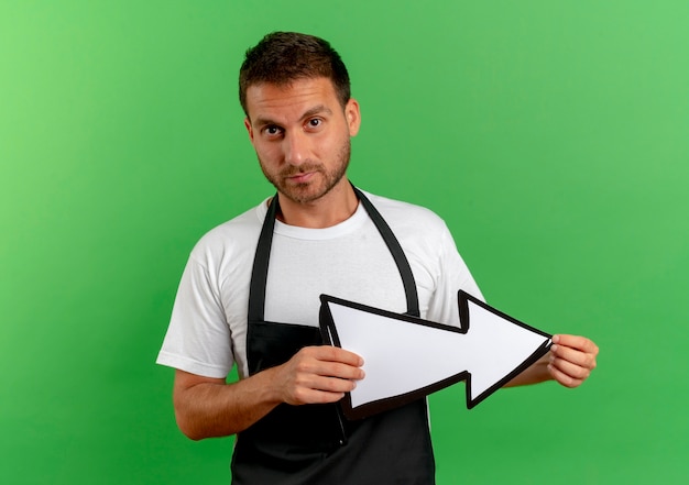 Foto gratuita uomo del barbiere in grembiule che tiene freccia bianca guardando in avanti con la faccia seria in piedi sopra la parete verde