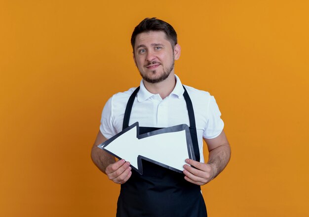 barber man in apron holding white arrow  with smile on face standing over orange wall