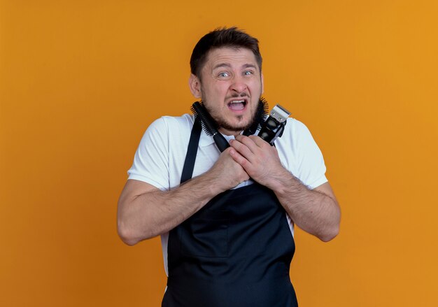 barber man in apron hugging beard trimmer and hair brushes  scarted and confused standing over orange wall