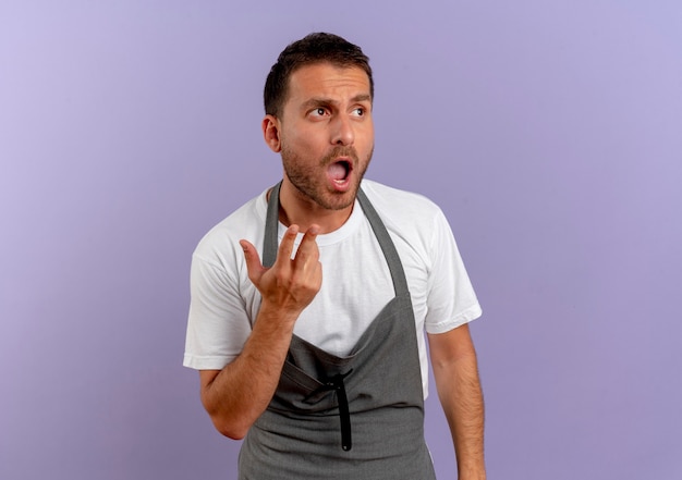 Barber man in apron looking aside confused with arm out as asking or arguing standing over purple wall