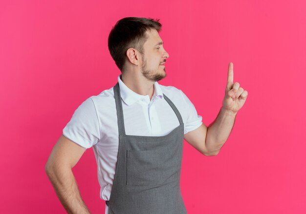 ピンクの壁の上に立って人差し指の笑顔を見せて脇を見てエプロンの床屋の男