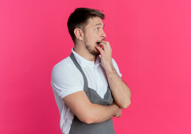 barber man in apron looking aside stressed and nervous biting nails standing over pink wall