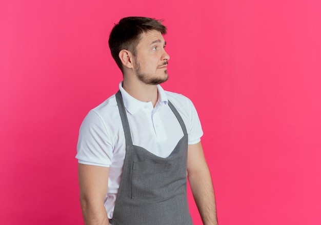 barber man in apron looking aside with sad expression standing over pink wall