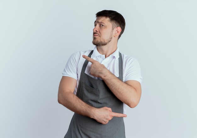 Barber man in apron looking aside with serious face pointing with index fingers to different directions standing over white background