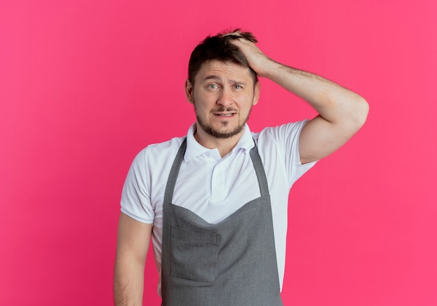 Barber man in apron looking at camera confused scratching head standing over pink background