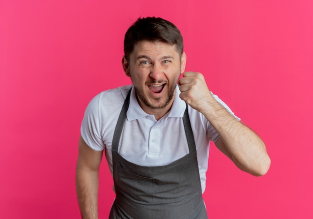 Free photo barber man in apron looking at camera going wild clenching fist shouting with aggressive expression standing over pink background