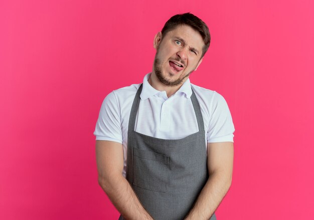 Barber man in apron looking at camera making grimace sticking out tongue standing over pink background