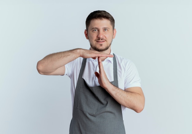 白い背景の上に立っている手でタイムアウトジェスチャーを作るカメラを見ているエプロンの理髪店の男