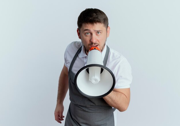 Barber man in apron looking at camera shouting through megaphone standing over white background