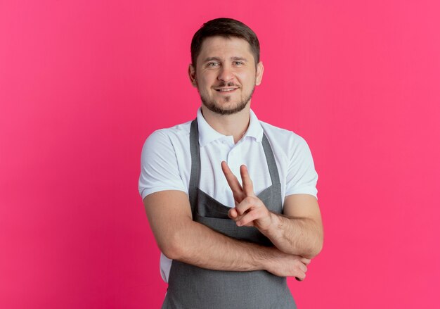 Barber man in apron looking at camera smiling showing victory sign standing over pink background