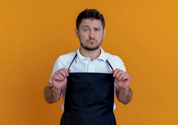 Barber man in apron looking at camera with confident expression standing over orange background