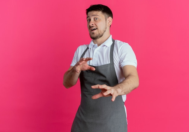 Barber man in apron looking at camera with disgusted expression making defense gesture with hands standing over pink background