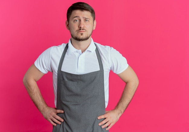 Foto gratuita barbiere uomo in grembiule che guarda l'obbiettivo con seria espressione fiduciosa in piedi su sfondo rosa