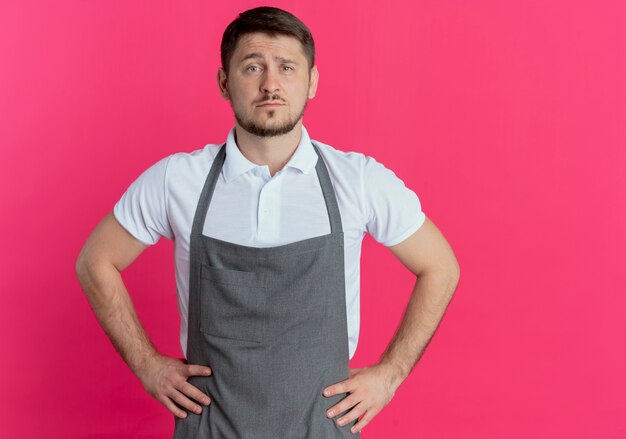 Barber man in apron looking at camera with serious confident expression standing over pink background