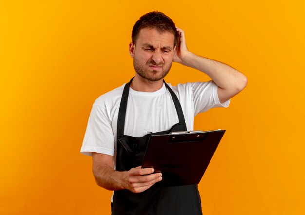 Barber man in apron looking at clipboard with confused expression standing over orange wall