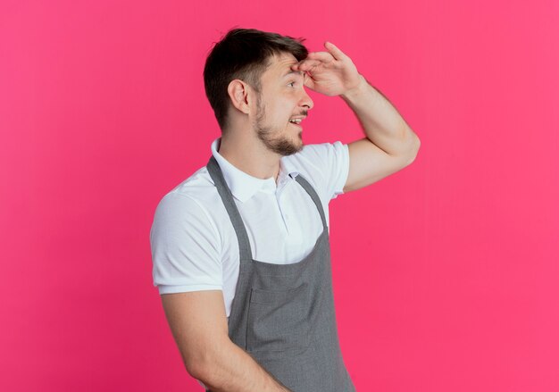 Free photo barber man in apron looking far away with hand over head to look something standing over pink wall