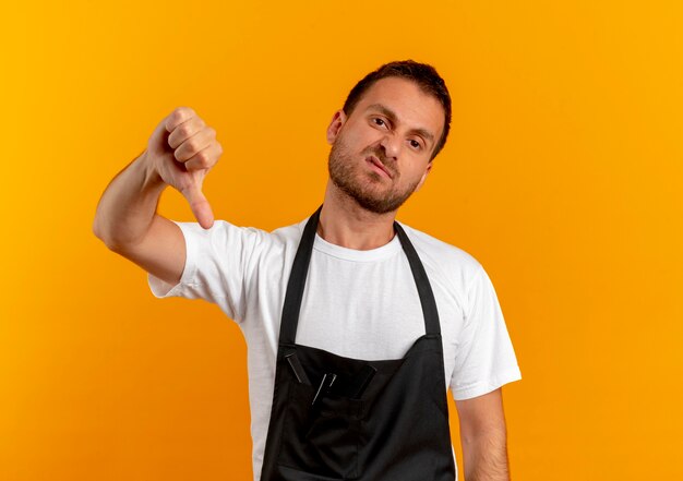 Barber man in apron looking to the front displeased showing thumbs down standing over orange wall