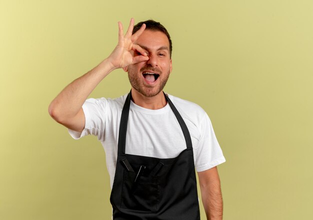 Barber man in apron looking to the front doing ok sign with fingers looking through fingers standing over light wall