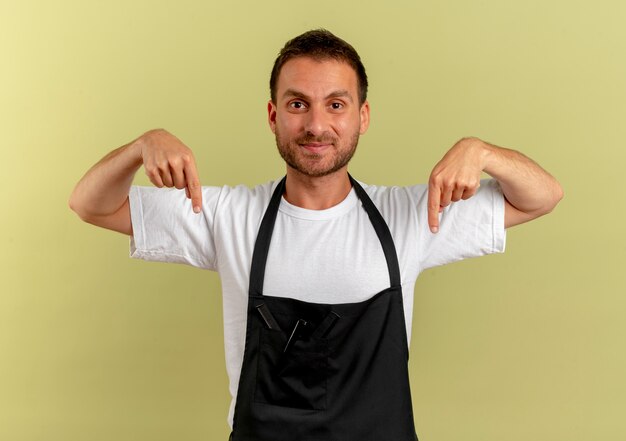 Barber man in apron looking to the front pointing with index fingers down standing over light wall