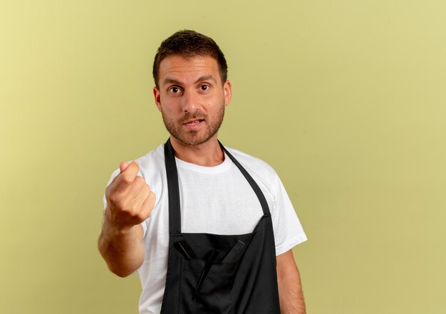 Barber man in apron looking to the front rubbing fingers asking for money standing over light wall