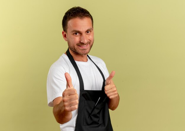 Barber man in apron looking to the front smiling cheerfully showing thumbs up standing over light wall