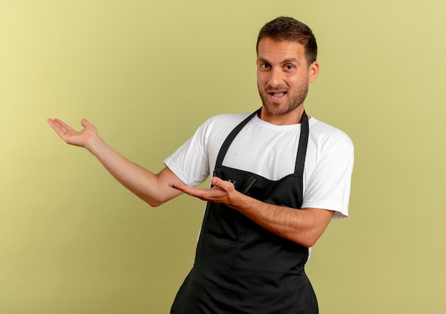 Barber man in apron looking to the front smiling presenting something with arms of his hands standing over light wall