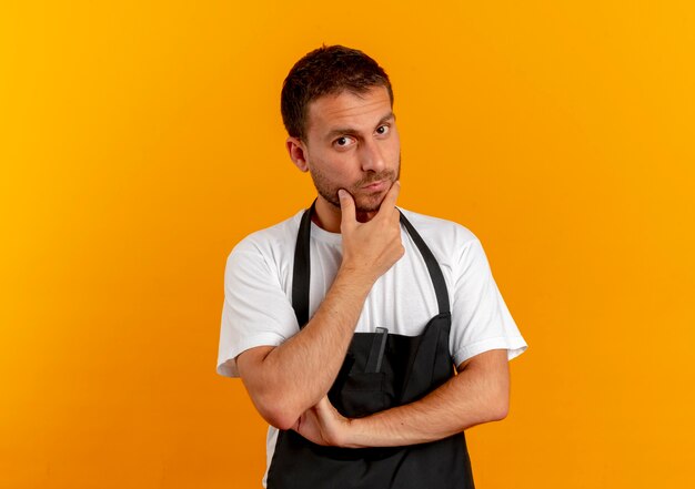 Barber man in apron looking to the front with hand on chin thinking standing over orange wall