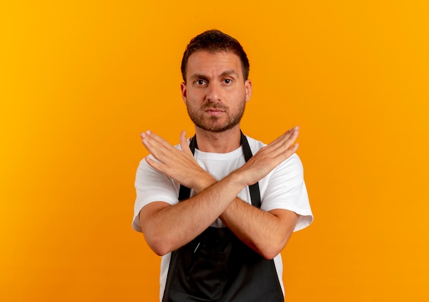 Barber man in apron looking to the front with serious face making stop gesture crossing hands standing over orange wall