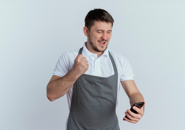 Barber man in apron looking at screen of his smartphone clenching fist happy and excited standing over white background