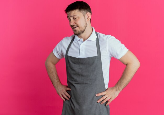 barber man in apron looking tiredand bored with arms at hip standing over pink wall