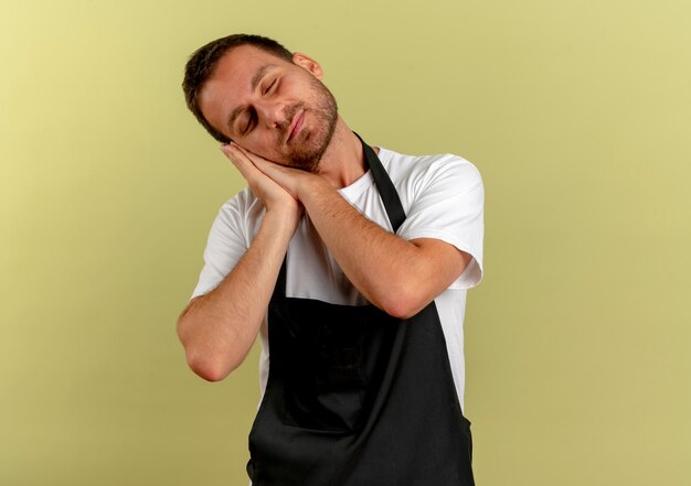 Barber man in apron making sleep gesture holding palms together standing over light wall