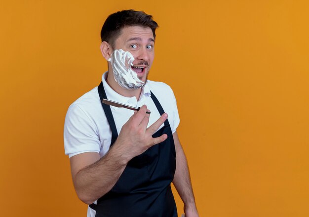 barber man in apron putting shaving foam on his beard  smiling showing razor standing over orange wall