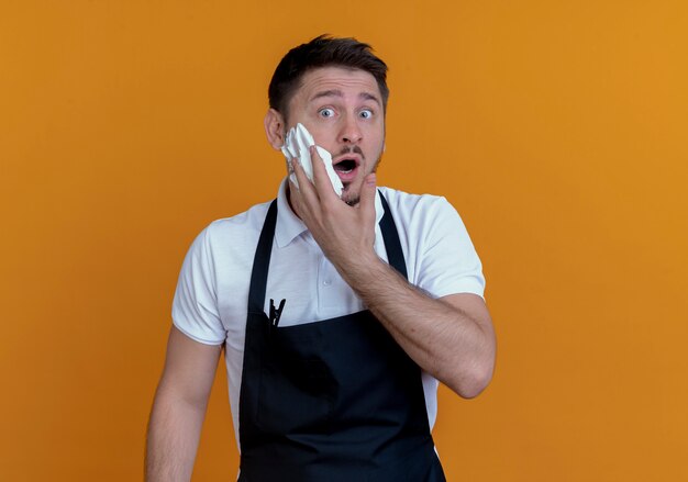 barber man in apron putting shaving foam on his beard  surprised standing over orange wall