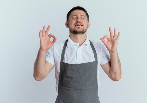 白い背景の上に立って目を閉じて指で瞑想ジェスチャーを作るリラックスエプロンの理髪店の男