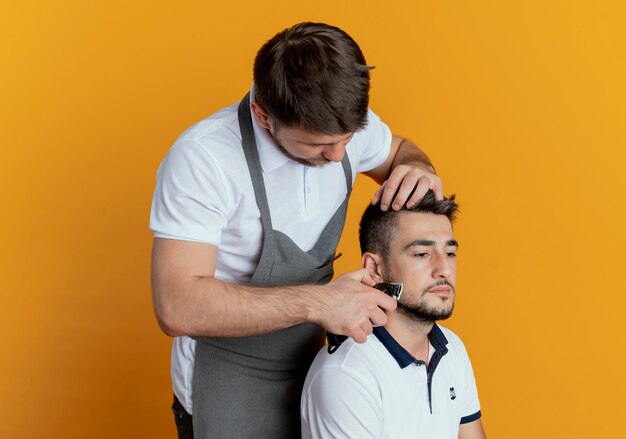 парикмахер в фартуке бреет бороду триммером для бороды довольного клиента над оранжевой стеной