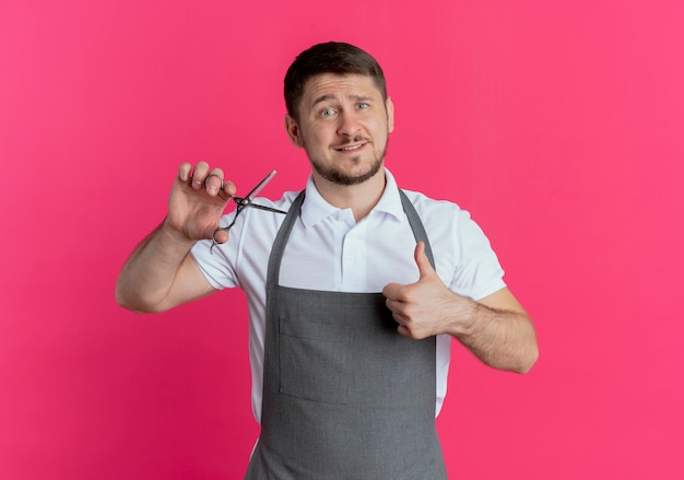 Barber man in apron showing scissors showing thumbs up standing over pink background